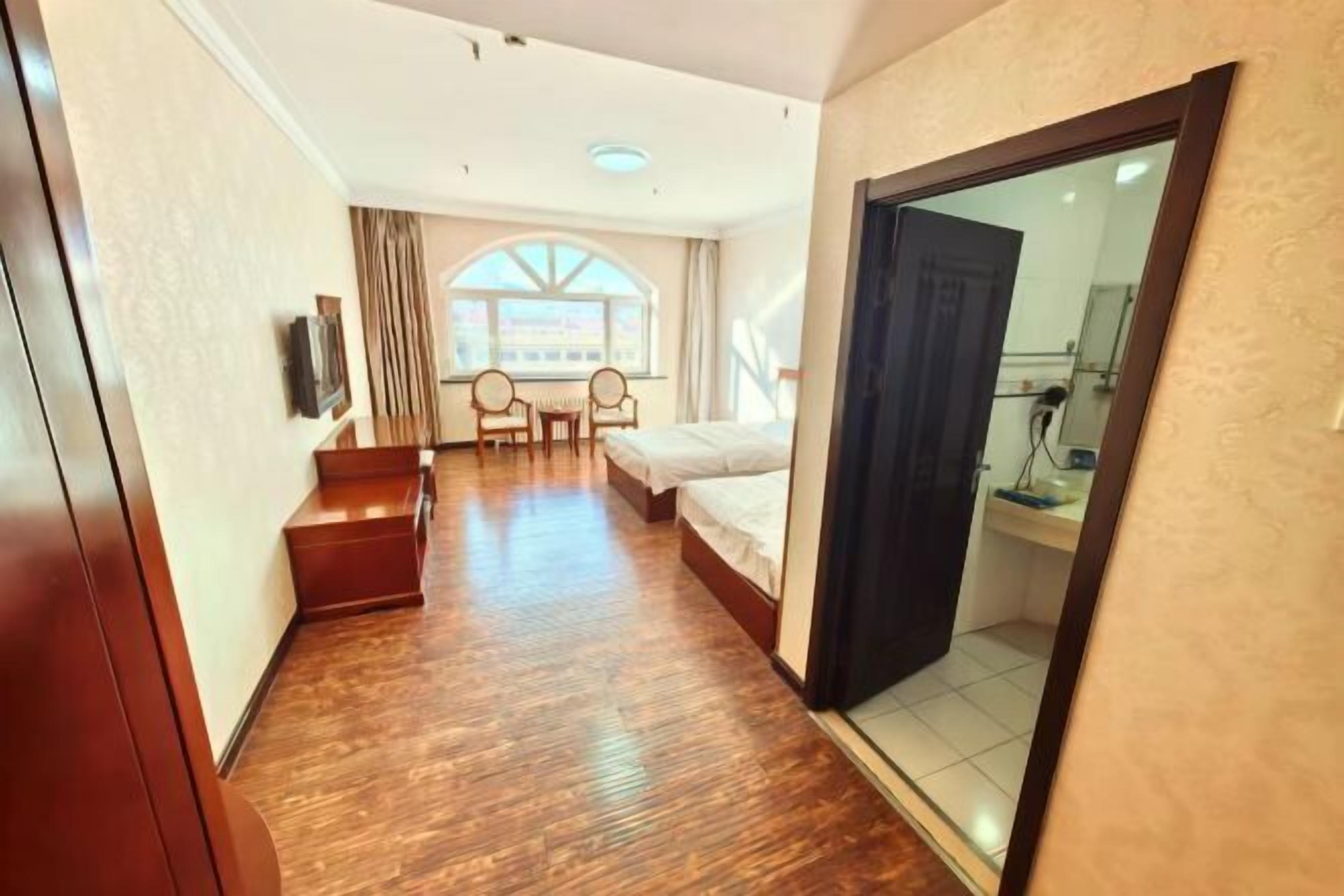 A hotel room with a wooden floor, a bed, a desk with a chair, a window with curtains, and a door.