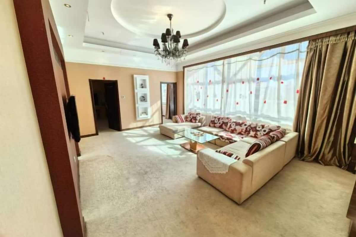 A spacious living room with a sofa set, a chandelier, and a large window with curtains.