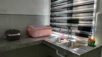 A kitchen with a sink, a pink storage bin, a rice cooker, and a window with horizontal blinds.
