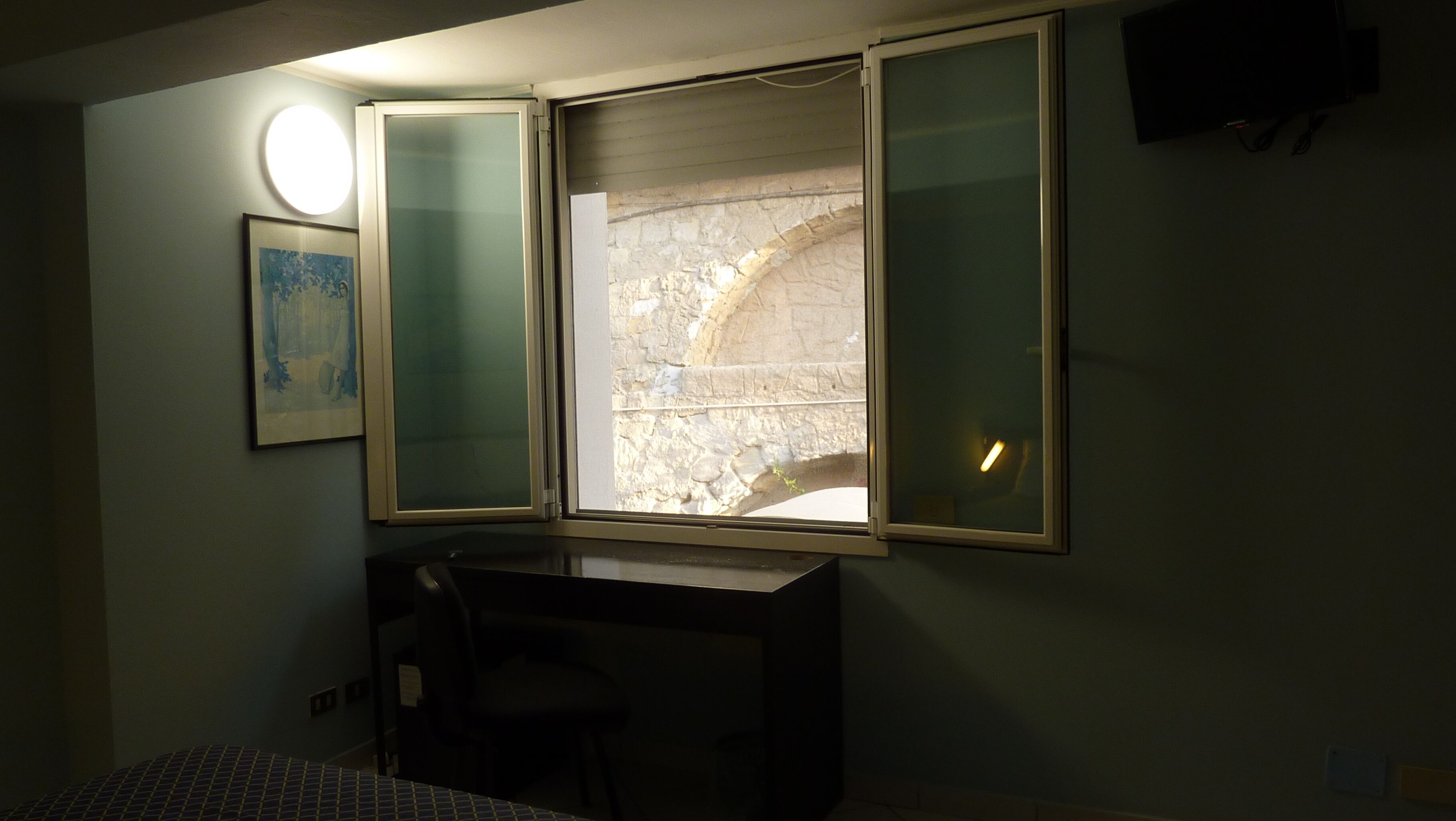 A room with a desk, a chair, a window with a view of stone walls, and a framed picture on the wall.