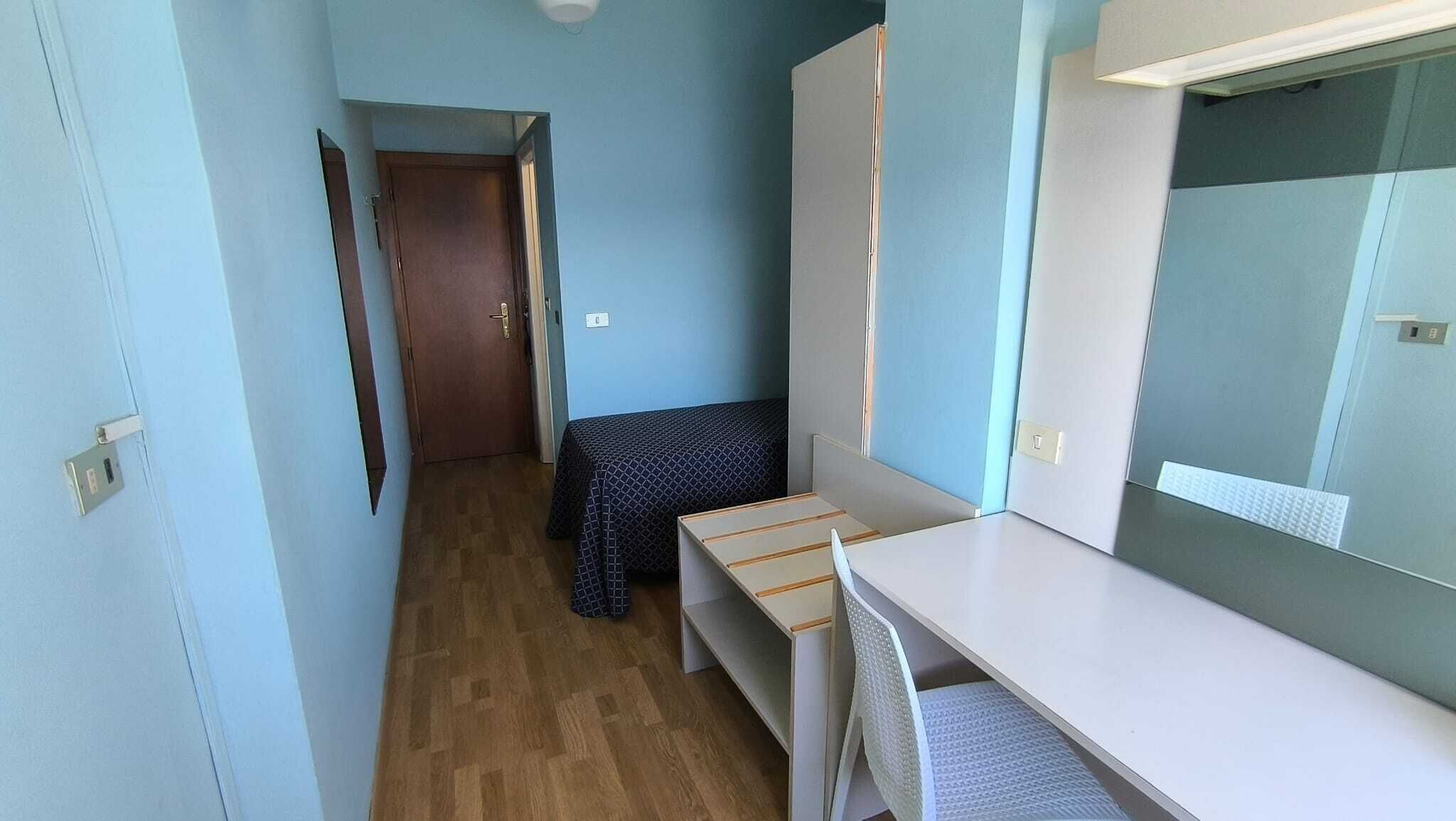 A narrow hallway with a wooden floor, a bed with a patterned bedspread, a desk with a chair, and a large mirror.
