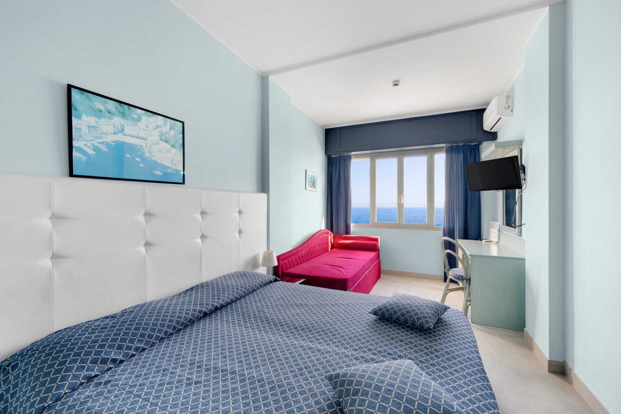 A hotel room with a large bed, a red sofa, a desk with a chair, and a TV.