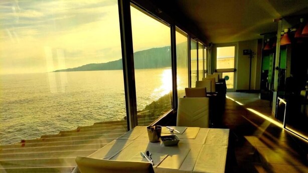 A dining area with a table set for two, large windows offering a view of the sea and distant land, and a modern interior design. - La Perla del capo hotel (Ospedaletti)