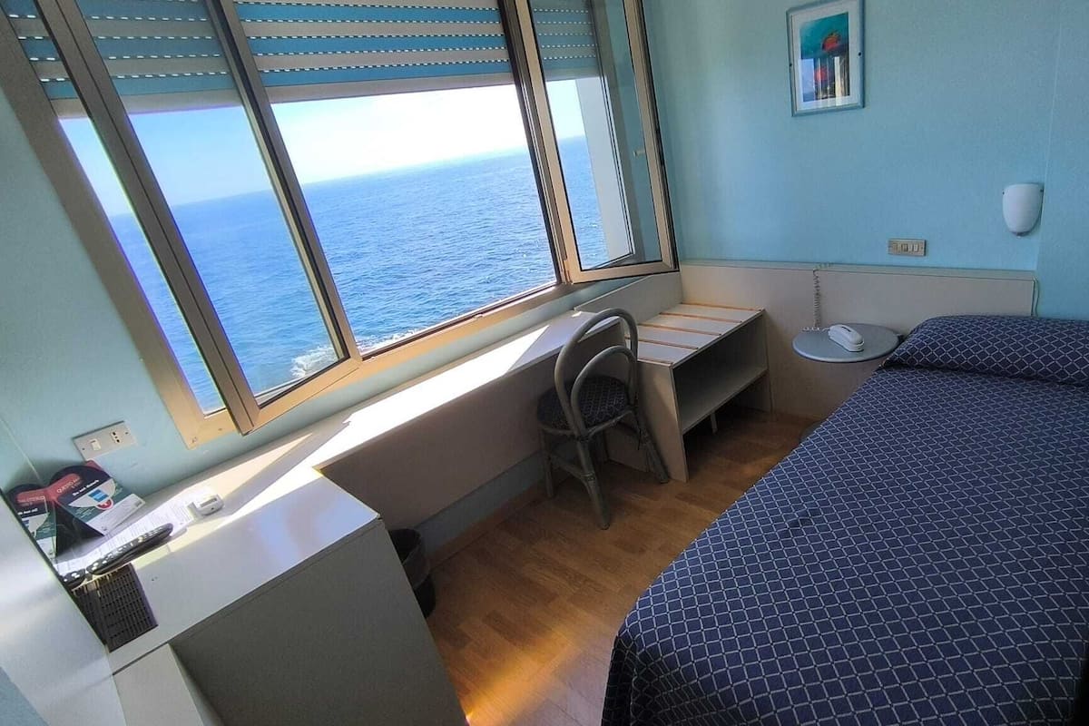 A compact room with a bed, a desk, and a chair. There is a window offering a view of the sea.