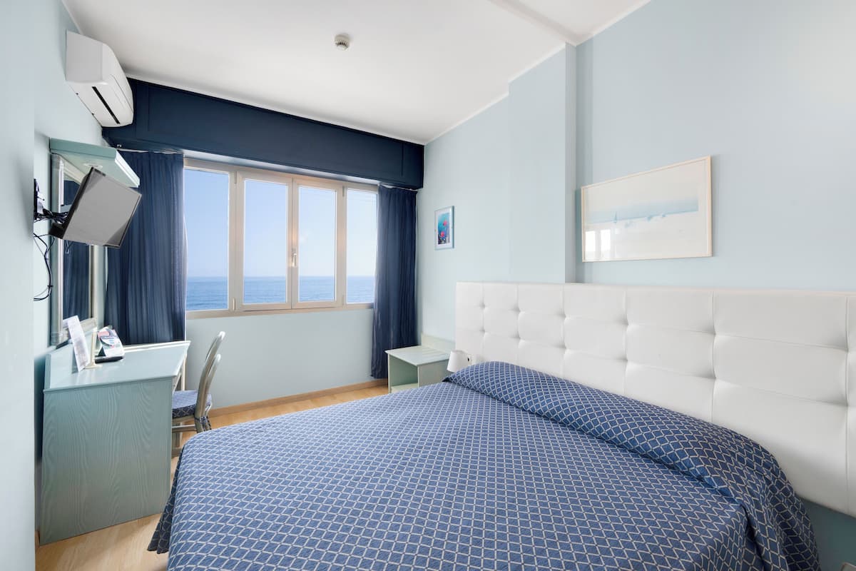 A hotel room with a bed, a desk, a chair, a nightstand, a window with curtains, and a wall-mounted air conditioner.