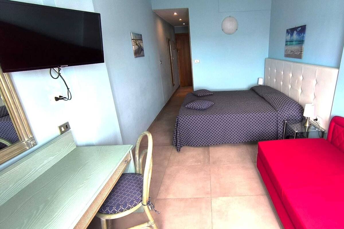 A small, compact room with a bed, a desk, and a chair. There is a television mounted on the wall and a picture frame on the wall.