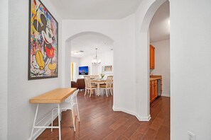 A dining area with a wooden table and chairs, a large Mickey Mouse painting on the wall, and an archway leading to a kitchen with wooden cabinets.