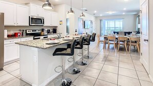 Una cocina moderna con armarios blancos, encimera de granito y electrodomésticos de acero inoxidable. Hay un comedor con mesa y sillas, y un ventanal con vistas.
