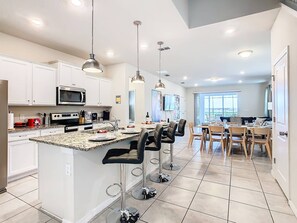 A modern kitchen with white cabinets, a granite countertop, and stainless steel appliances. There is a dining area with a table and chairs, and a large window offering a view.