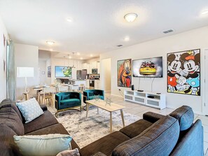 A modern living room with a sofa, a coffee table, and a TV. There is a kitchen in the background with cabinets and a microwave.