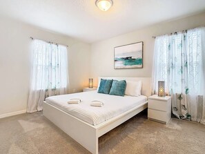 A bedroom with a large bed, two bedside tables, a painting above the bed, and sheer curtains with floral patterns.