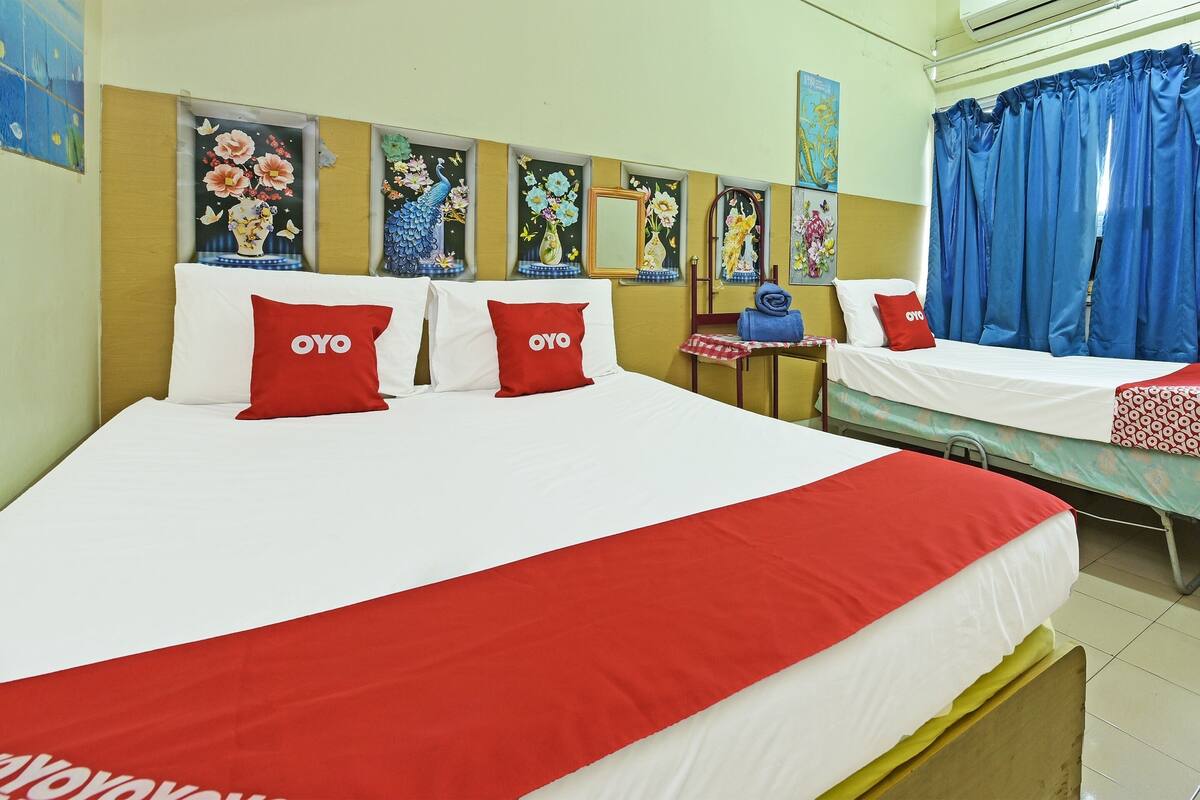 Quarto de hotel com duas camas, cada uma com roupa de cama branca e vermelha, e almofadas vermelhas com o logotipo "OYO". Há quadros florais na parede, uma pequena mesa com uma cadeira e cortinas azuis.
