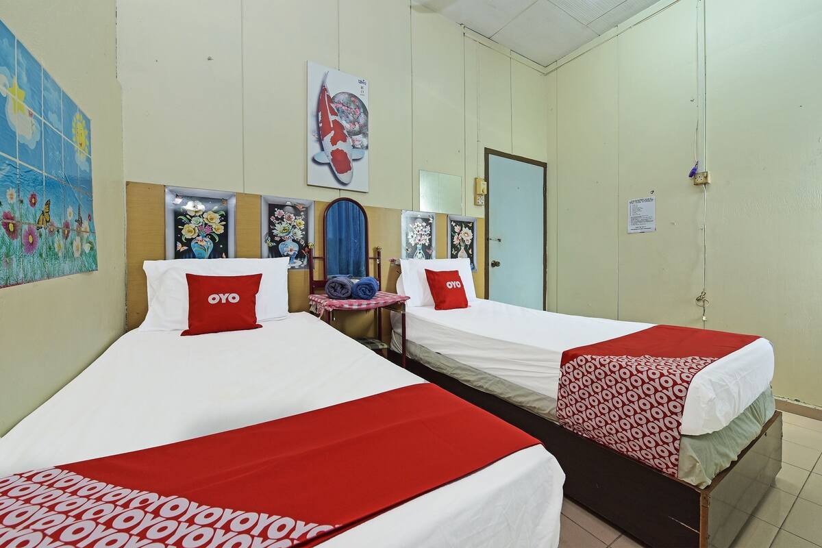 Quarto de hotel com duas camas, cada uma com colcha e travesseiros vermelhos e brancos, uma mesinha com abajur e obras de arte nas paredes.
