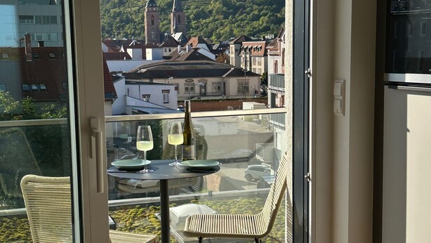 A balcony with a table and chairs, a bottle of wine, and a view of a town with buildings and a hill. - Modern city apartment (Neustadt an der Weinstraße)