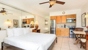 Un dormitorio con una cama grande, ventilador de techo y kitchenette con armarios de madera y electrodomésticos modernos.