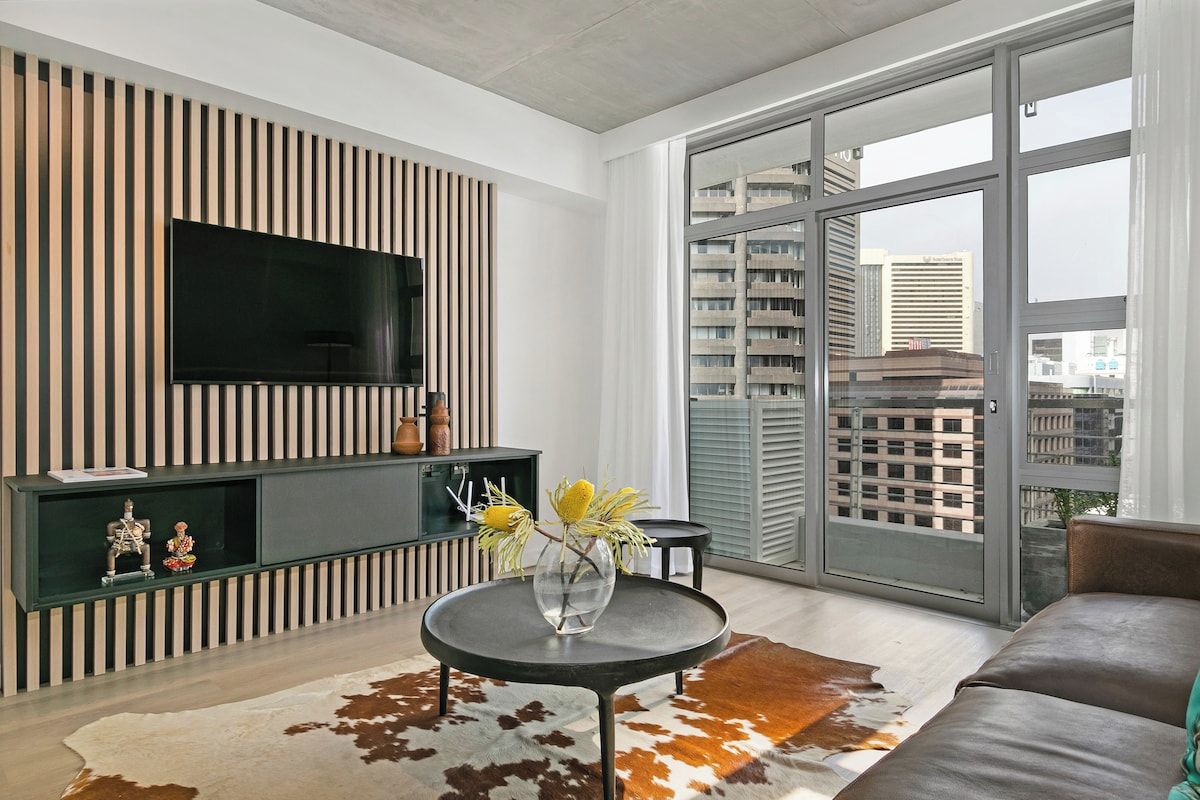 A modern living room with a flat-screen TV mounted on a wall, a round coffee table with a vase, and a large window offering a city view.