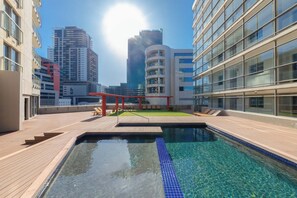 A rooftop swimming pool with a wooden deck, surrounded by modern buildings and a clear sky.