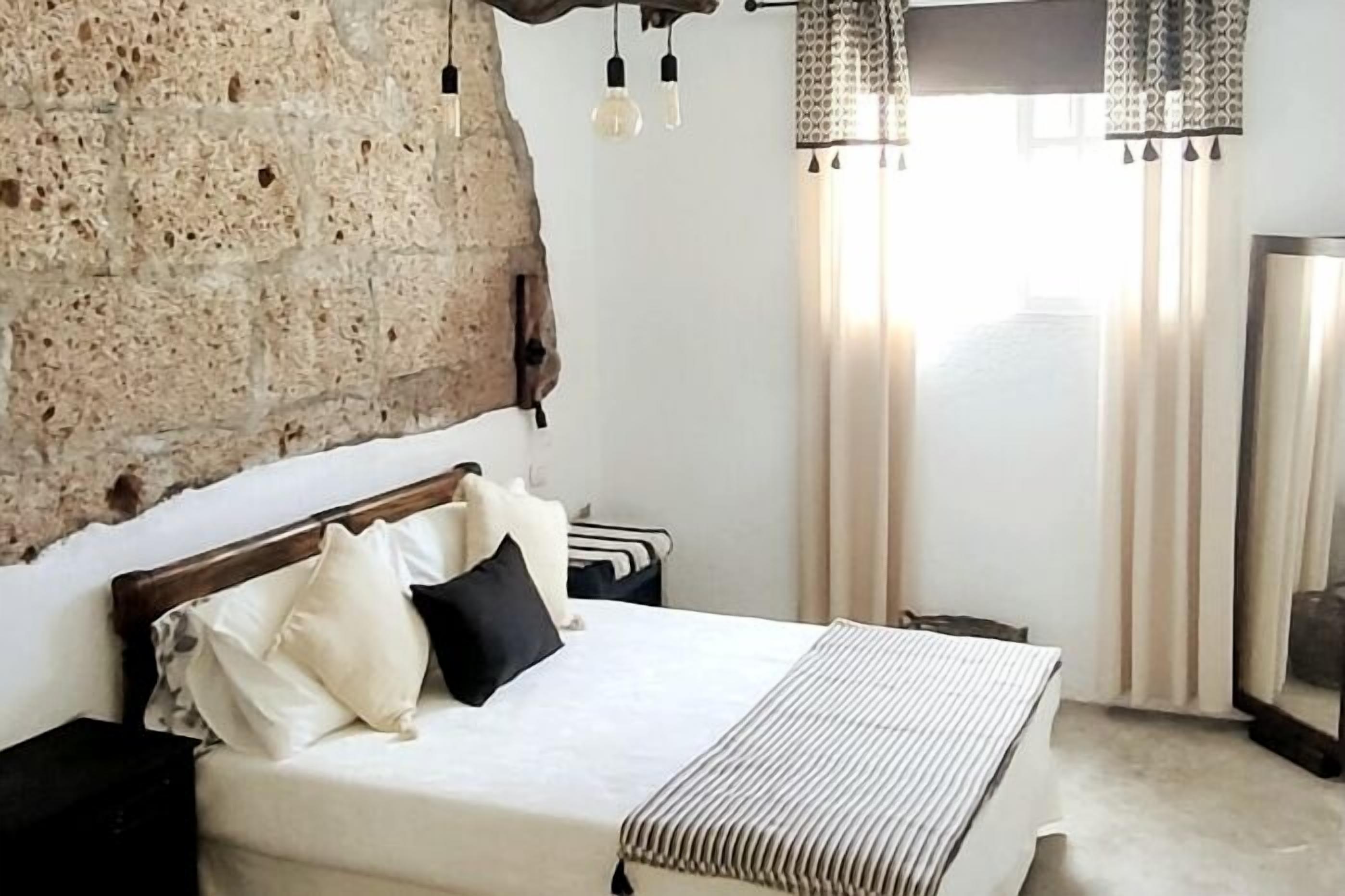 Un dormitorio con pared de piedra, una cama con ropa blanca y almohadas negras, una mesita de noche y una lámpara colgante.