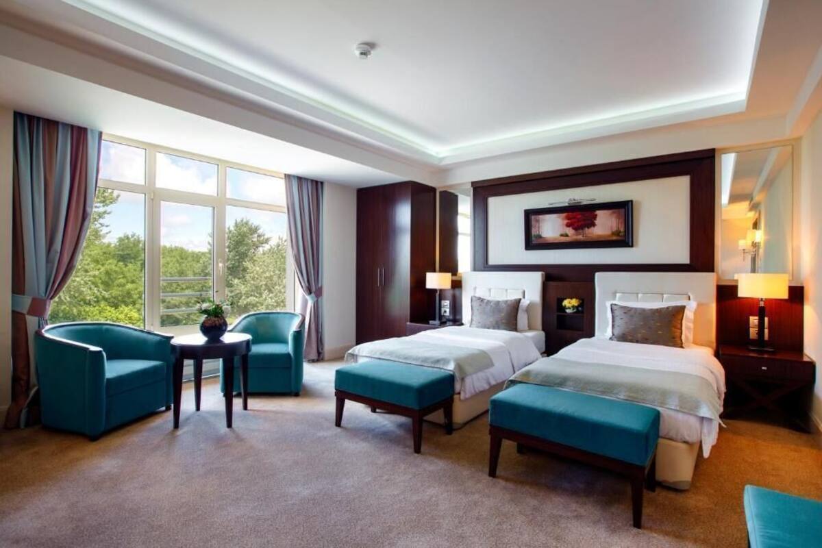 A hotel room with two beds, a desk, chairs, and a large window with a view.