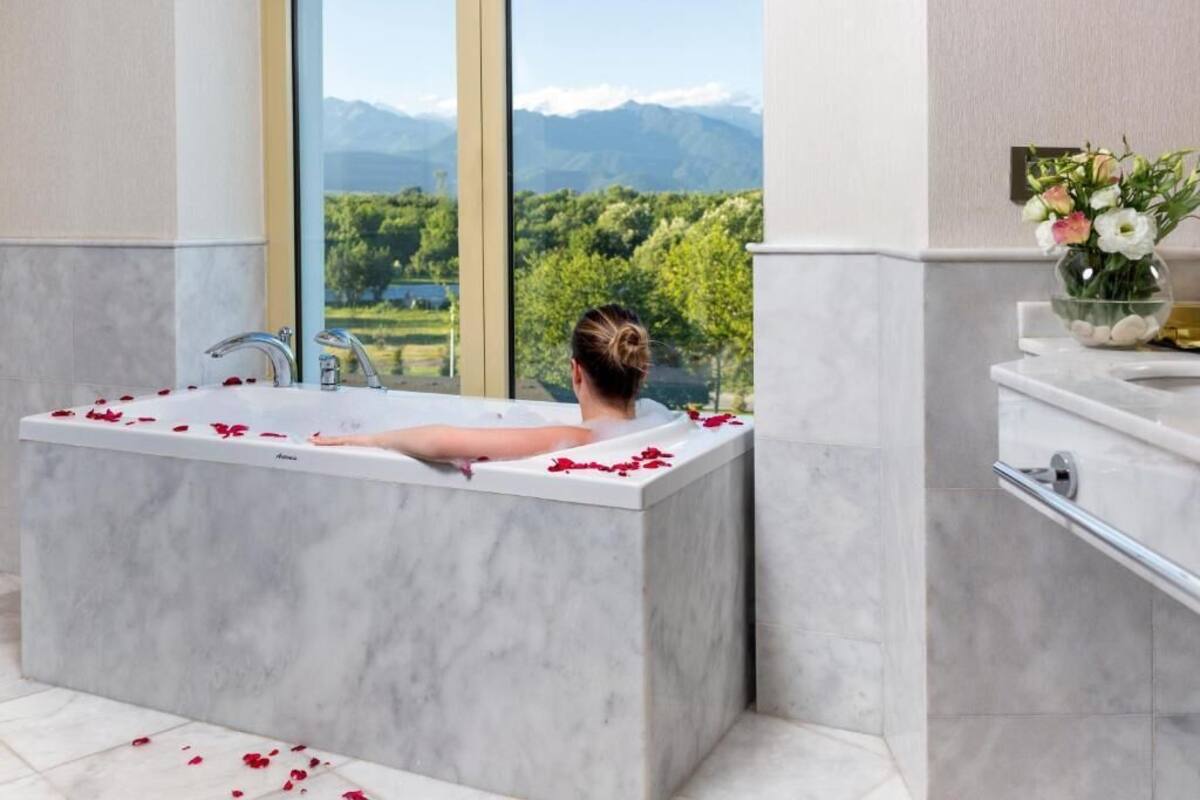 A modern bathroom with a large bathtub, marble walls, and a view of mountains through a large window.