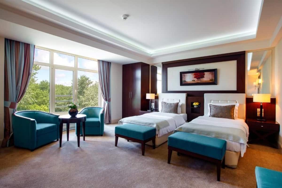 A hotel room with two beds, a sofa, a small table, and a view of trees through large windows.