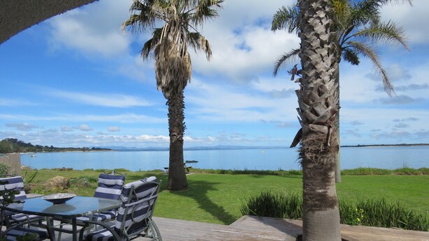 An outdoor deck with a table and chairs, overlooking a body of water with palm trees and a distant shoreline.
