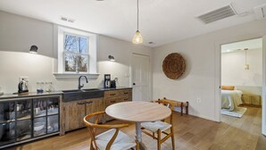 A modern kitchen with a round wooden table, wooden chairs, a built-in cabinet with glass doors, a window, and a woven wall hanging.