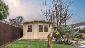Una baita di legno con tetto spiovente, un cortile recintato e un piccolo albero con un secchio appeso.