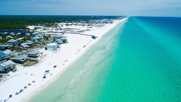 Vue aérienne d’un complexe hôtelier en bord de mer, avec de nombreux bâtiments, une longue étendue de sable blanc et une eau turquoise cristalline.