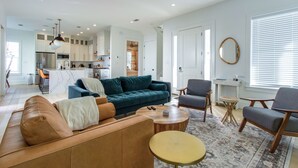 Un salon moderne avec un canapé, des fauteuils et une table basse. Au fond, on aperçoit une cuisine équipée de placards et d’un réfrigérateur.