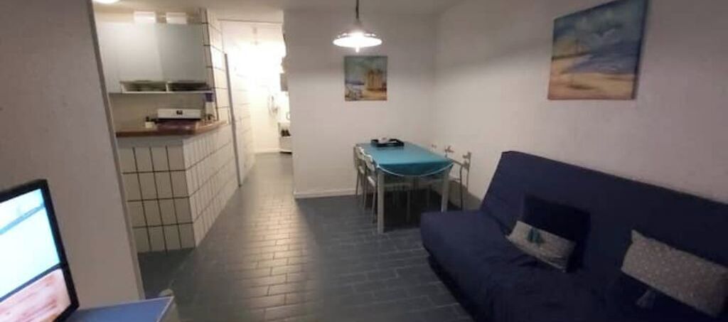 A narrow hallway leading to a dining area with a table and chairs, a blue sofa, and a kitchen area with cabinets and a countertop.