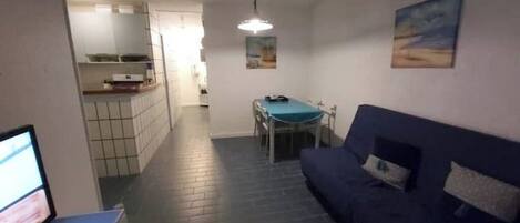 A narrow hallway leading to a dining area with a table and chairs, a blue sofa, and a kitchen area with cabinets and a countertop.