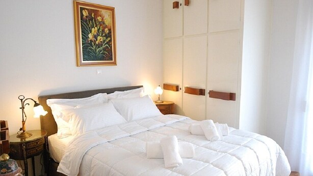 Een net opgemaakt bed met witte lakens en handdoeken, een bedlampje, een decoratieve vaas, en een schilderij van bloemen aan de muur.