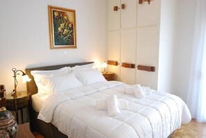 Una cama bien hecha con ropa de cama y toallas blancas, una lámpara de noche, un jarrón decorativo y un cuadro de flores en la pared.