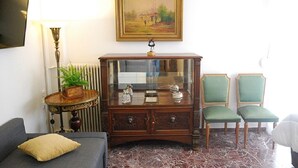 Een woonkamer met een bank, een houten kast met glazen deuren, twee groene fauteuils, een lamp en een schilderij aan de muur.