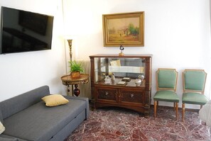 Un salon avec un canapé gris, un meuble en bois doté de portes en verre, un tableau accroché au mur et un téléviseur fixé au-dessus du canapé.