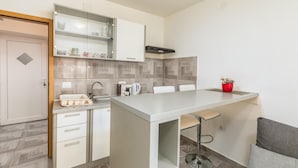 Een compacte keuken met een geïntegreerd baronderdeel, een spoelbak en een fornuis. Er is een klein zithoekje met een stoel en een tafel.