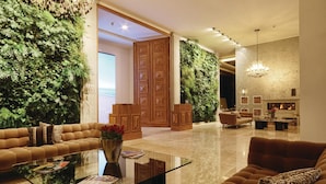 Een ruime lobby met een grote bank, een glazen bijzettafeltje en een open haard. De muren zijn versierd met weelderige groene begroeiing, en er is een houten deur die naar een andere kamer leidt.
