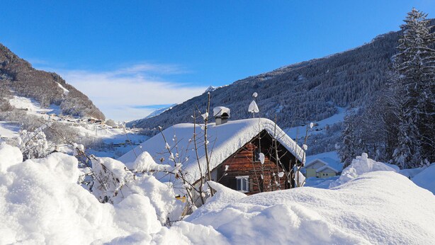 Une cabane en bois, dont le toit est recouvert de neige, entourée d’un paysage enneigé et de montagnes.