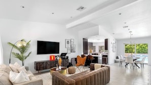 Een moderne woonkamer met een grote flatscreen-tv, een bank, een eethoek met uitzicht op het zwembad, en een keuken op de achtergrond.