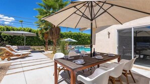 Een terras met een houten tafel, witte stoelen en een grote parasol.