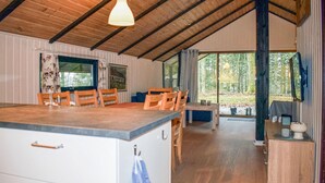 Ein Interieur einer Holzhütte mit einem Holztisch, Stühlen, einer Küchenzeile mit Schränken und einem großen Fenster mit Blick auf den Wald.