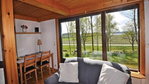 Een knus interieur met een houten plafond, een grijze bank met witte kussens, een houten eettafel met stoelen, en grote ramen met uitzicht op een groen landschap.