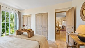 Ein Schlafzimmer mit einem großen Bett, einem Korbtisch mit Tablett und Blick auf einen Garten durch ein großes Fenster.