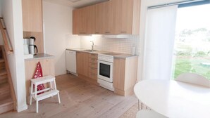 A compact kitchen with wooden cabinets, a dishwasher, an oven, and a sink. There's a small white stool and a window with a view of greenery. - Awesome Home in Burgsvik With 3 Bedrooms and Wifi (Burgsvik)