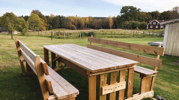 Una mesa de picnic de madera con bancos en un campo cubierto de césped, con una casa al fondo.