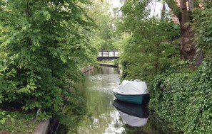 A narrow canal with a boat docked on the right, surrounded by dense greenery and a wooden bridge in the background.