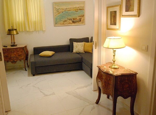 Un salon avec un canapé gris, une petite table d’appoint en bois sur laquelle se trouve une lampe, un tableau accroché au mur et un sol en marbre.