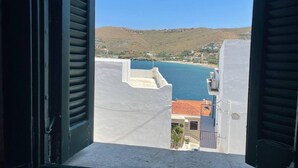 A view from a window with shutters open, overlooking a coastal area with buildings, a hill, and a body of water. - Charming two Bedroom Fisherman's House on Kea (Kea)