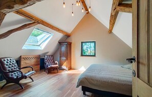 Un dormitorio acogedor en el desván, con techo inclinado, vigas de madera, un ventanal grande, una cama, una silla y un armario.